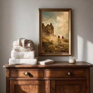 Puede incluir: Una impresi&oacute;n de arte mural vertical enmarcada que representa una ruina de piedra en una colina con dos figuras, colocada sobre una c&oacute;moda de madera junto a una pila de toallas blancas dobladas y un peque&ntilde;o portavelas de cristal.