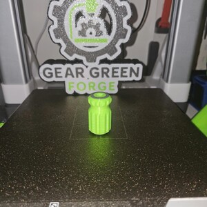 May include: A vibrant green, cylindrical 3D-printed object with a ridged body and a dark grey top ring. The object is on a black surface, with a "GEAR GREEN FORGE" logo in the background.