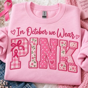 Peut inclure: Un sweat-shirt à col rond rose clair avec le texte In October we Wear PINK en écriture rose foncé. Le mot PINK est affiché en grandes lettres majuscules avec des motifs variés incluant du vichy rose, des fleurs et des pois.