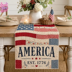 May include: A patriotic table runner with a patchwork design featuring red, white, and blue fabrics. The word "AMERICA" is prominently displayed, along with "EST. 1776". The runner is decorated with stars, floral patterns, and a heart. A small American flag and floral arrangement are also present.