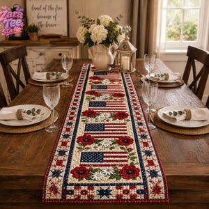 May include: A rectangular fabric table runner featuring a repeating pattern of American flags interspersed with red poppies and white daisies. The runner has a decorative border with blue stars and red squares on a cream background, laid on a table.