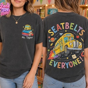 Op de afbeelding: Een antracietgrijs t-shirt met een kleine afbeelding op de borst van een gele schoolbus op een stapel boeken met de tekst Ms. Taylor. De achterkant toont een grote gele schoolbusafbeelding met de tekst SEATBELTS EVERYONE!