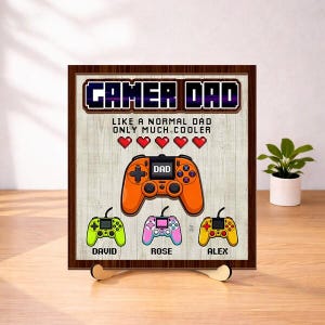 May include: A wooden framed sign with the words "GAMER DAD" in a pixelated font. The sign features an orange game controller with the word "DAD" on it, and three smaller controllers below in green, pink, and yellow. The names "David", "Rose", and "Alex" are below the controllers.