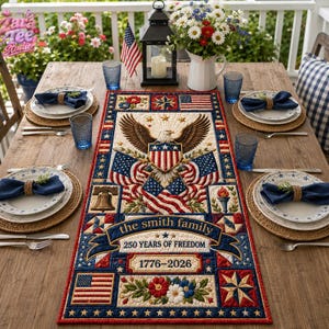 May include: A rectangular fabric table runner featuring a central bald eagle with an American flag shield, surrounded by patriotic motifs, the text 'the smith family', '250 YEARS OF FREEDOM', and '1776-2026', set on a wooden table with place settings.