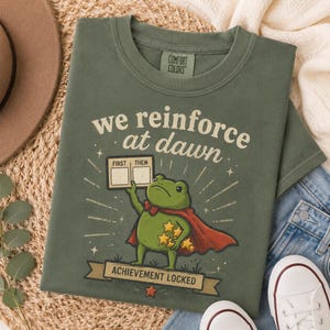 May include: An olive green t-shirt featuring a graphic of a green frog wearing a red cape and bow tie. The frog holds a sign labeled "FIRST THEN" and a cluster of yellow stars. Text reads "we reinforce at dawn" and "ACHIEVEMENT LOCKED" on a banner.