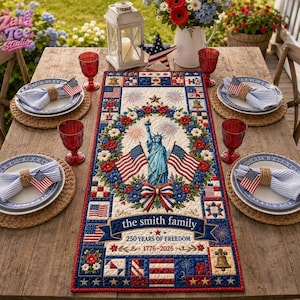 May include: A rectangular table runner featuring a patriotic design with the Statue of Liberty, American flags, and floral accents. The text reads 'the smith family', '250 YEARS OF FREEDOM', and '1776-2026'. It is set on a wooden table with plates, napkins, and red glasses.