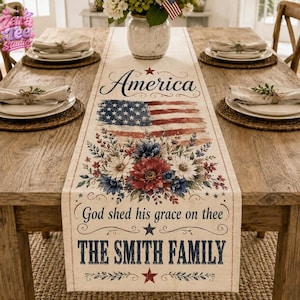 Puede incluir: Camino de mesa rectangular beige con diseño de bandera americana y detalles florales. Las palabras "America", "God shed his grace on thee" y "THE SMITH FAMILY" están impresas. Se encuentra sobre una mesa de madera, lista para una comida.