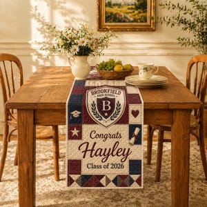 May include: A quilted table runner with a graduation theme. The runner features a central crest with the text "Brookfield High School" and "B". Below the crest, the text reads "Congrats Hayley Class of 2026". The runner is in shades of red, white, and blue.