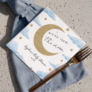 May include: A square white paper napkin featuring a gold glitter crescent moon, light blue watercolor clouds, and small gold stars. The text reads "we're over the moon" and "Sophia's Baby Shower 06.22.XX" in a black script and sans-serif font.