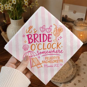 May include: A square paper napkin with a light pink and white vertical striped pattern. The text reads it's BRIDE O'CLOCK Somewhere, FELICITY'S BACHELORETTE, and Miami FL. Graphics include heart-shaped sunglasses, a cocktail, a seashell, and a beach chair.