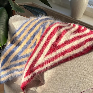 Può includere: Uno scialle a righe fatto a mano nei toni del rosso, bianco, blu e giallo è appoggiato su una sedia dai toni neutri. Il design dello scialle presenta un motivo a spina di pesce. L'immagine include anche una pianta a foglie grandi e un vaso di fiori secchi.