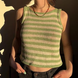 Könnte beinhalten: Ein grün-weiß gestreiftes Tanktop. Das ärmellose Oberteil ist aus Strickmaterial gefertigt. Das Model trägt eine Perlenkette und eine dunkle Jeans. Der Schatten des Models wird auf die Wand geworfen.