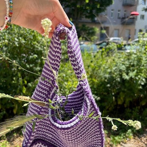 Puede incluir: Un pequeño bolso tejido a mano, morado y blanco, con un patrón de chevrón. El bolso tiene un ribete blanco y está lleno de vegetación. El bolso se sujeta por un asa hecha del mismo material tejido.