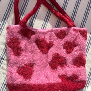 Könnte beinhalten: Handgefertigte rosa Tragetasche mit einem Muster aus roten Herzen. Die Tasche hat rote Griffe und einen roten Boden. Die Tasche besteht aus einem weichen, strukturierten Material, ideal für den täglichen Gebrauch.
