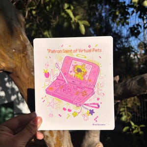 May include: A pink illustration featuring a handheld gaming device with a cartoon character inside. The text "Patron Saint of Virtual Pets" is at the top. The image has a whimsical style with stars, hearts, and other decorative elements.