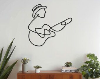 May include: Black metal wall art depicting a musician playing a guitar. The artwork is a continuous line drawing of a person wearing a hat and playing a guitar. The art is mounted on a white wall above a wooden cabinet.
