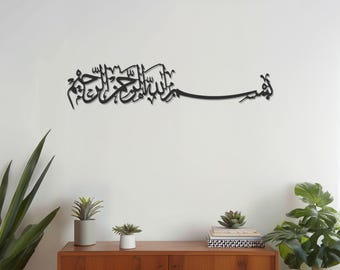 May include: Black metal wall art featuring Arabic calligraphy. The artwork is mounted on a white wall above a wooden cabinet with plants. The calligraphy is a flowing script, adding a touch of elegance to the space.