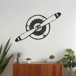 May include: A monochrome metal wall art piece featuring an aeroplane propeller. The propeller has two blades with white tips and is mounted on a circular engine housing. The artwork is mounted on a white wall.