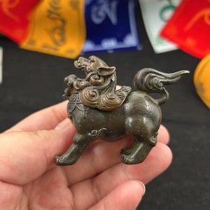 Puede incluir: Una figura de bronce de una criatura mítica, posiblemente un león o un perro, con detalles intrincados. La escultura tiene una melena detallada, una cola rizada y se sostiene en una mano. El fondo incluye banderas de oración coloridas.