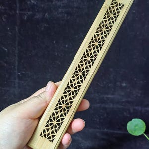 以下が含まれることがあります： 上部に装飾的な花の模様が彫られた長方形の竹製お香立て。ホルダーは薄いタン色で、人の手で持たれています。背景は暗く、質感のある表面です。