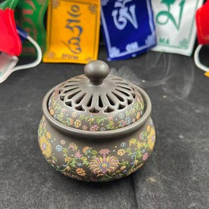 Może przedstawiać: Ciemnobrązowe ceramiczne kadzielnica o okrągłym kształcie z pokrywką. Powierzchnię zdobi kolorowy, misterny wzór kwiatowy z żółtymi, różowymi, niebieskimi i zielonymi pnączami. Pokrywka posiada centralny uchwyt oraz ozdobne otwory wentylacyjne dla dymu.