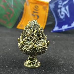 May include: A detailed bronze incense burner with a flame-like design. The burner sits on a pedestal base with intricate carvings. Colorful prayer flags are visible in the background, adding a spiritual element to the scene.