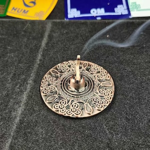Könnte beinhalten: Ein bronzefarbener Räucherstäbchenhalter mit floralem Design. Der runde Halter hat eine zentrale Tasse mit einem vertikalen Pfosten und ist von aufwendigen Mustern umgeben. Rauch steigt vom Halter auf, was auf die Verwendung hindeutet.