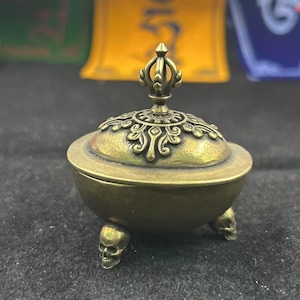 May include: A bronze-colored incense burner with a decorative lid and three skull-shaped feet. The lid is adorned with intricate patterns and a stylized finial. The burner has a round base and a detailed design, suitable for spiritual practices.