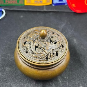 Op de afbeelding: Een bronzen wierookbrander met een gedetailleerd, opengewerkt deksel met een feniksontwerp. De ronde brander heeft een kleine knop aan de bovenkant en geeft een wolkje rook af. De algehele kleur is een warm, antiek brons.