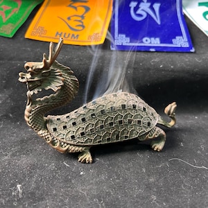 May include: A bronze dragon-shaped incense burner with a turtle-like body. The dragon has detailed scales and a fierce expression. Smoke rises from the burner, and colorful prayer flags are in the background. The burner is approximately 15 cm long.
