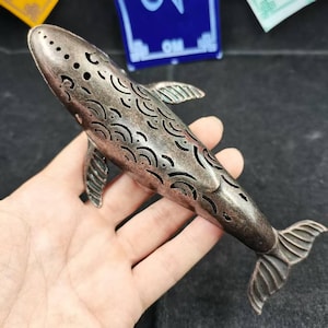 May include: A bronze-coloured whale-shaped incense burner. The whale has cut-out wave patterns along its body and several small holes on top. The fins and tail are detailed. The burner is held in a hand.