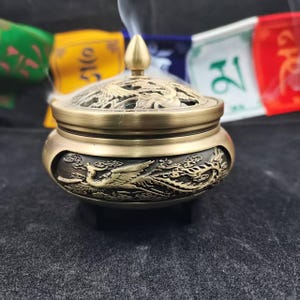 Op de afbeelding: Een bronzen wierookbrander met ingewikkelde draken- en wolkenreliëfs. De deksel heeft een opengewerkt ontwerp en een conische finial. Er stijgt rook op uit de brander. Op de achtergrond zijn kleurrijke gebedsvlaggen te zien.