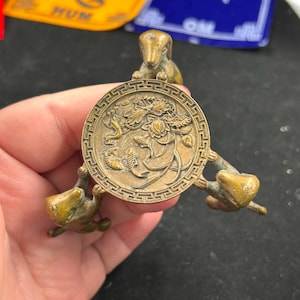 Pode incluir: Um suporte decorativo de três pernas na cor bronze. O suporte apresenta um topo circular com um desenho em relevo de flores e peixes, sustentado por três figuras em forma de cão. O suporte é segurado em uma mão.