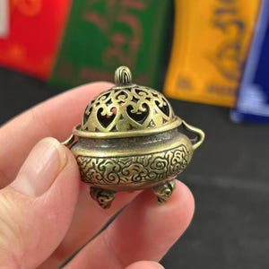 Puede incluir: Un pequeño quemador de incienso de latón antiguo con detalles intrincados. El quemador presenta una tapa abovedada con recortes en forma de corazón y patrones decorativos alrededor de la base. Tiene tres patas pequeñas y dos asas.
