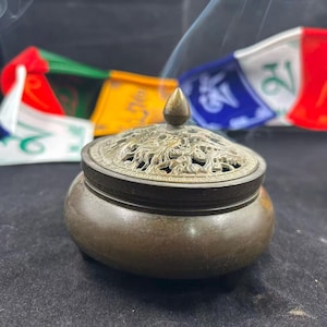 Op de afbeelding: Een bronzen wierookbrander met een decoratief, opengewerkt deksel en een kleine, afgeronde behuizing. Het deksel is bekroond met een kleine, puntige finial. Wispeltjes rook stijgen op uit de brander en kleurrijke gebedsvlaggen zijn op de achtergrond.