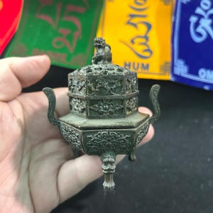 Puede incluir: Un quemador de incienso de bronce detallado con intrincados patrones florales y geométricos. El quemador tiene una pequeña figura de león en la parte superior y asas curvas. La base tiene tres patas decorativas. El fondo incluye banderas de oración coloridas.