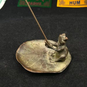 May include: A bronze incense holder featuring a frog figurine seated on a lily pad. The frog holds an incense stick, and the lily pad has a detailed leaf pattern. The holder is designed to catch ash.