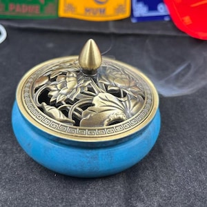 Op de afbeelding: Een blauwe wierookbrander met een decoratief bronzen deksel met een bloemenmotief. Het deksel heeft een kleine, puntige knop. Wispels rook stijgen op uit de brander, wat suggereert dat deze in gebruik is. De brander is rond en heeft een eenvoudig, elegant ontwerp.