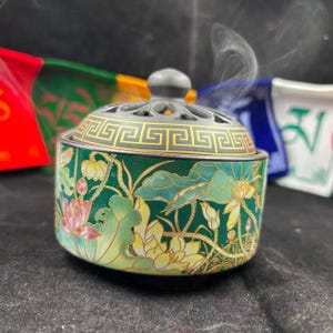 May include: A round ceramic incense burner with a dark green base featuring a colorful lotus flower and leaf pattern. It has a removable grey lid with a gold Greek key border and a perforated top, with visible wisps of smoke rising from it.