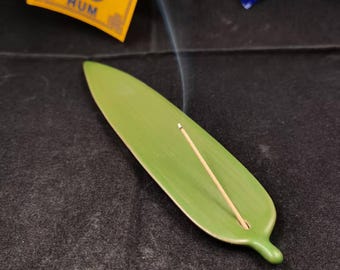 May include: A green ceramic leaf-shaped incense holder with a lit incense stick. The holder is on a dark surface, with wisps of smoke rising. A yellow tag with the word "HUM" is visible in the background.