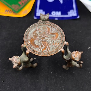 Op de afbeelding: Een bronzen decoratieve standaard met een ronde plaat met een drakenontwerp, ondersteund door drie katachtige figuren. De standaard heeft een antieke uitstraling en is waarschijnlijk van metaal.