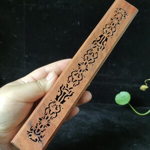 Peut inclure: Un porte-encens rectangulaire en bois, orné de motifs ajourés complexes de fleurs et de formes géométriques. Le porte-encens est d'une couleur brun rougeâtre chaleureuse, probablement en bois dur. Le motif est symétrique et s'étend sur toute la longueur.