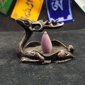 May include: A dark brown incense holder in the shape of a deer with a purple incense cone. The incense holder has a tray to catch the ash. The background includes colorful prayer flags.