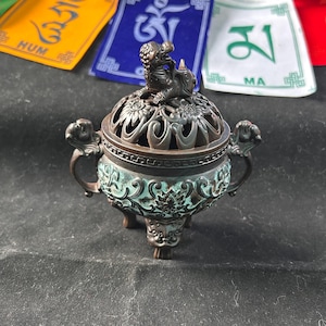 May include: A bronze incense burner with detailed floral and scrollwork. The burner features a small statue on the lid and handles. The base has a turquoise patina, and it stands on three small feet. Colorful prayer flags are in the background. The burner is a decorative item.