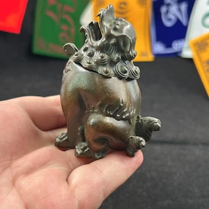 May include: A bronze incense burner sculpted as a mythical lion. The detailed figure has an open mouth, a curled mane, and a cub at its feet. The burner is held in a hand, with colourful prayer flags in the background.