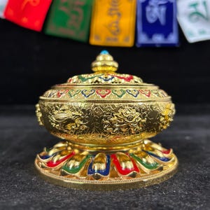 May include: A gold-colored metal bowl with a matching lid, featuring intricate embossed floral and scroll patterns. The base is decorated with a ring of petal-like shapes in red, green, and blue enamel accents, topped with a small blue bead.