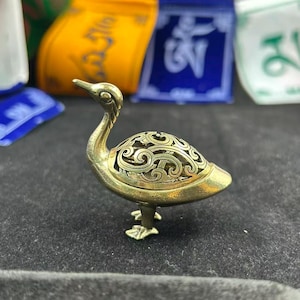 Puede incluir: Un quemador de incienso con forma de pato de latón con intrincados calados en su cuerpo. El pato tiene un cuello largo y se apoya sobre pequeños pies. El fondo incluye banderas de oración coloridas.