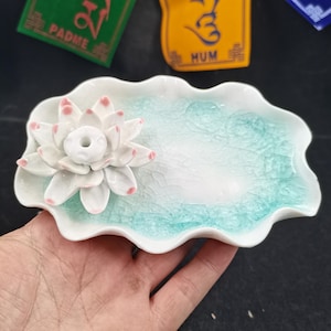 Puede incluir: Un porta incienso de flor de loto de cerámica blanca con un plato festoneado azul claro y blanco. La flor de loto tiene detalles en rosa en los pétalos. El plato tiene un acabado craquelado.