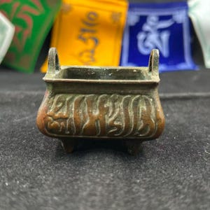 May include: A small, antique bronze incense burner with a square shape and ornate, embossed designs. It has two small handles and three feet. The burner is a dark bronze color with hints of green and brown.