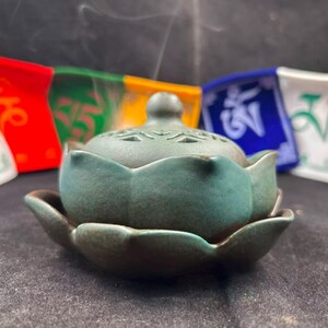 May include: A matte teal ceramic incense burner shaped like a blooming lotus flower with a removable lid. The lid features intricate cut-out patterns and a small knob handle. Wisps of smoke rise from the center of the burner, which is set against a backdrop of colorful prayer flags.